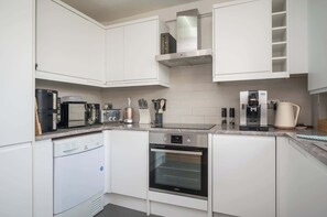 Una cocina moderna con muebles blancos, lavavajillas, horno, cafetera y hervidor.