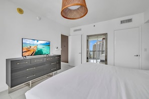 A bedroom with a bed, a TV on a dresser, a door, and a view of buildings.