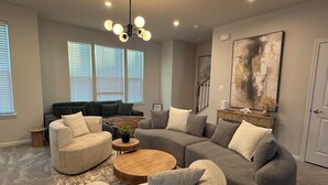 Una sala de estar moderna con un sofá seccional, dos sillones y una mesa de centro. Hay tres ventanas con persianas, un cuadro abstracto de gran tamaño y una escalera al fondo.