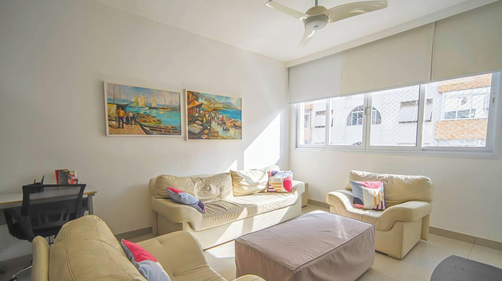 A living room with a sofa set, a coffee table, and two armchairs. There are two paintings on the wall, a ceiling fan, and a window with a view of buildings outside.