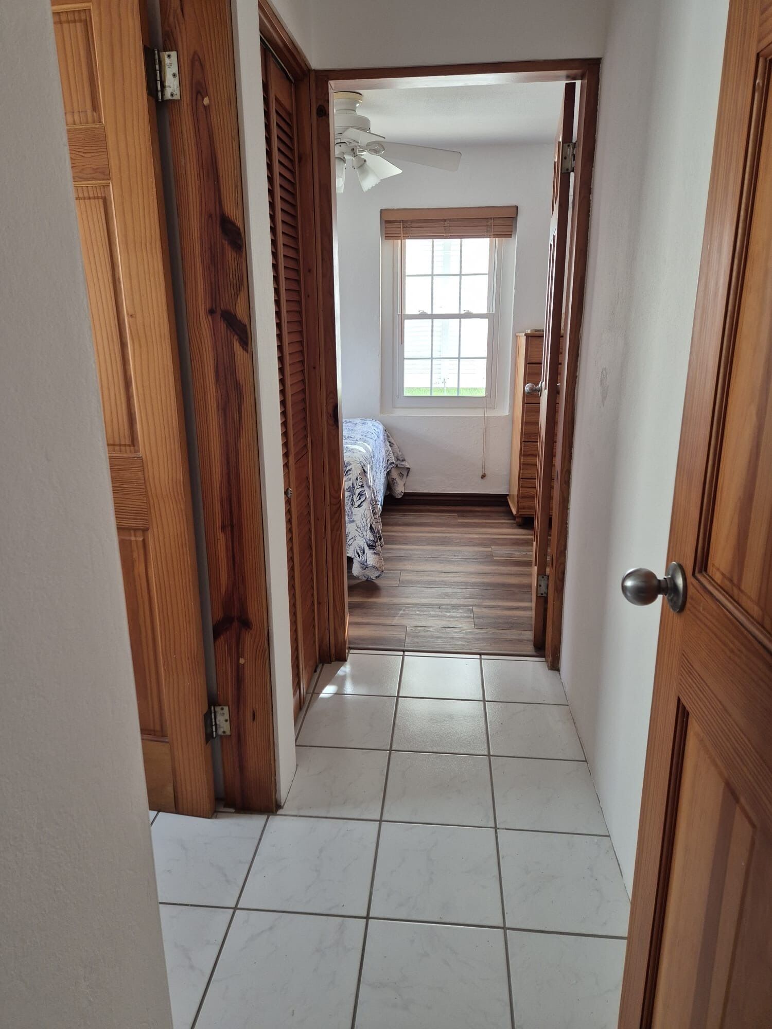 Un pasillo con puertas de madera, un ventilador de techo, una ventana con persianas y una cama con edredón estampado.
