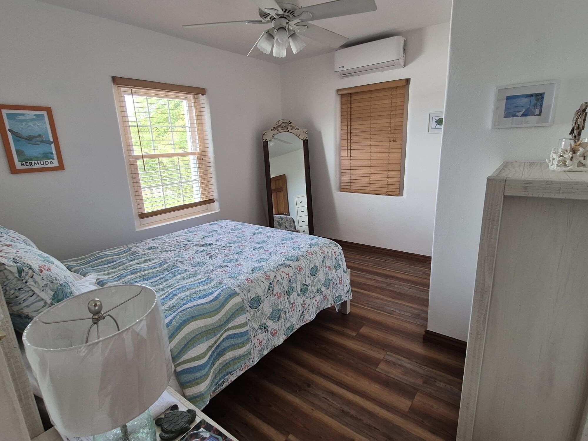 Une chambre à coucher comprenant un lit, un ventilateur de plafond, une fenêtre avec des stores, un miroir et une commode.