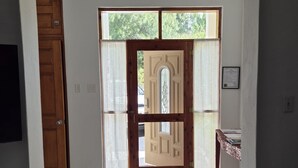 Un pasillo con suelo de baldosas, una puerta de madera, una puerta de cristal con un diseño decorativo y una pequeña mesa con mantel estampado.