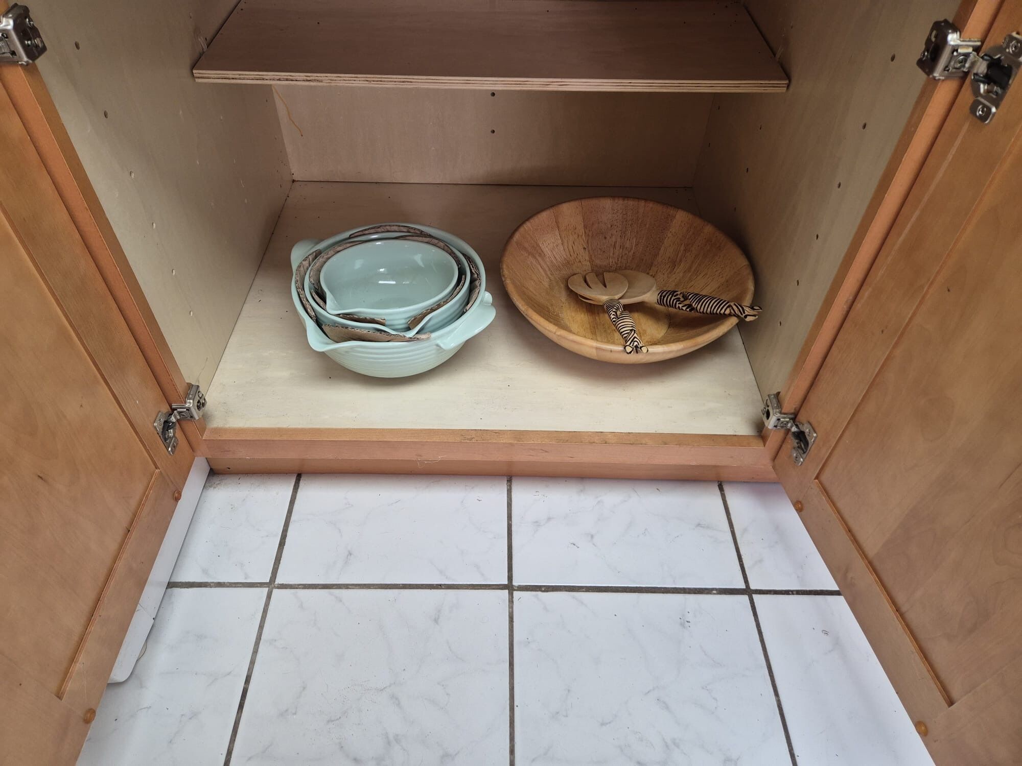 A kitchen cabinet with a marble countertop, containing a stack of plates and a wooden bowl with a spoon.