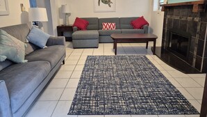 Una sala de estar con un sofá gris, un sofá gris más pequeño, una mesa de centro y un tapete estampado. Hay una chimenea con un revestimiento de azulejos y un cuadro de una tortuga en la pared.