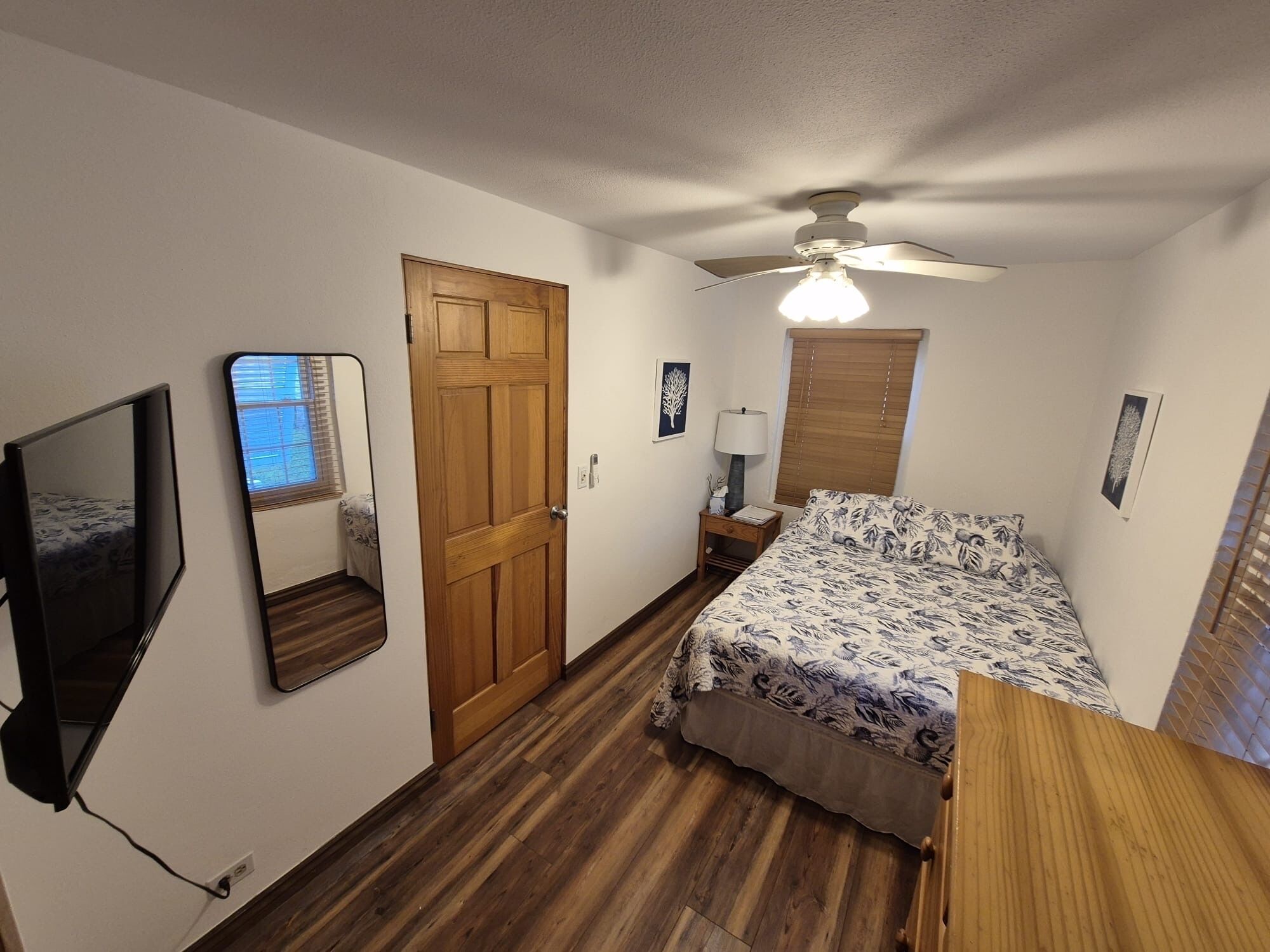 Een slaapkamer met een houten vloer, een bed met een bedspread met patroon, een plafondventilator, een houten deur, een nachtkastje met een lamp en een aan de muur bevestigde televisie.