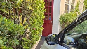Un coche blanco estacionado junto a un edificio con puerta roja, pared blanca y una cámara de seguridad.