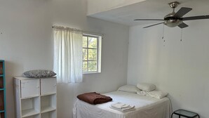 Un dormitorio con una cama individual, una ventana con cortinas blancas, un ventilador de techo y una mesita.