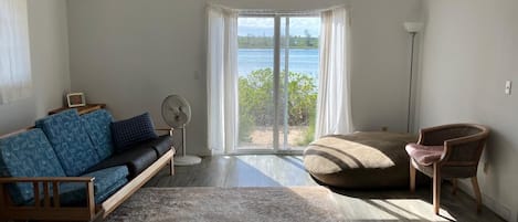 A spacious living room with a large window offering a view of the sea, a ceiling fan, a sofa, a chair, and a small table.