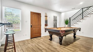 Een kamer met een pooltafel, een houten deur, een trap en een raam met jaloezieën.