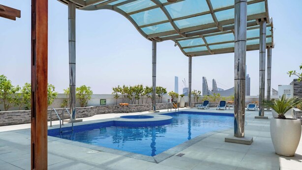 An outdoor swimming pool with a glass roof, surrounded by lounge chairs and potted plants, set against a city skyline.