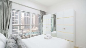 A hotel room with a large bed, a wardrobe with gold accents, a balcony with a view of tall buildings, and a window with curtains.