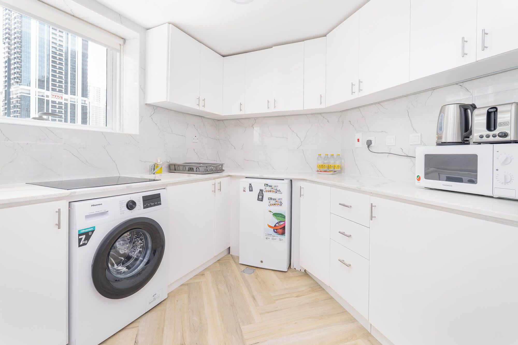 A compact kitchen with white cabinets, a built-in washing machine, a microwave, and a kettle.