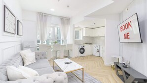 A modern apartment with a living area, a dining area, a kitchen, and a laundry room. There is a 'BOOK ME' sign on the wall.