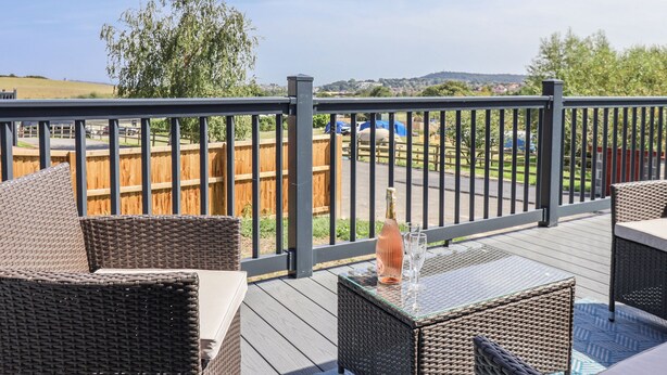 A deck with wicker furniture and a glass table, overlooking a fenced yard and distant hills.