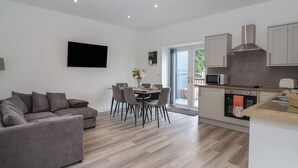 A modern living room with a grey sofa, dining area with chairs, a flat-screen TV mounted on the wall, and a kitchen with cabinets and a microwave.