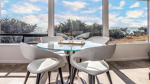 Un espace repas moderne avec une table en verre et des chaises blanches, de grandes fenêtres offrant une vue sur l’océan et les arbres.