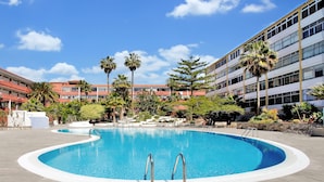 A swimming pool with a curved edge, surrounded by a paved area with palm trees and a multi-story building in the background. - 'Chasna' Holidays with Shared Pool, Shared Garden and Wi-Fi (Arona)