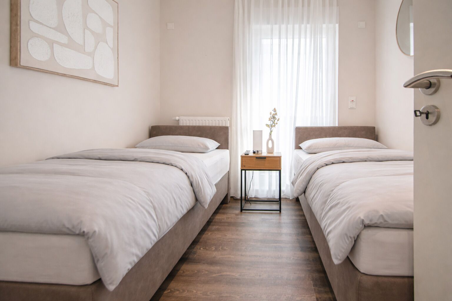 Et hotelværelse med to enkeltsenge, et natbord med en vase, en radiator og et vindue med let gardin.