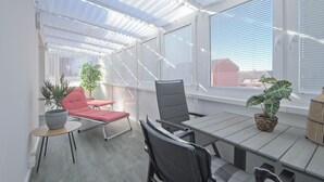 A sunlit patio with a table and chairs, large windows with blinds, and potted plants. - Your exclusive penthouse in a perfect location - pure luxury and relaxation with its own (Norderney)