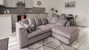 Un salon moderne avec un canapé d’angle gris, un coin repas avec une table et des chaises, une cuisine équipée de placards et d’une cuisinière, et une horloge au mur.