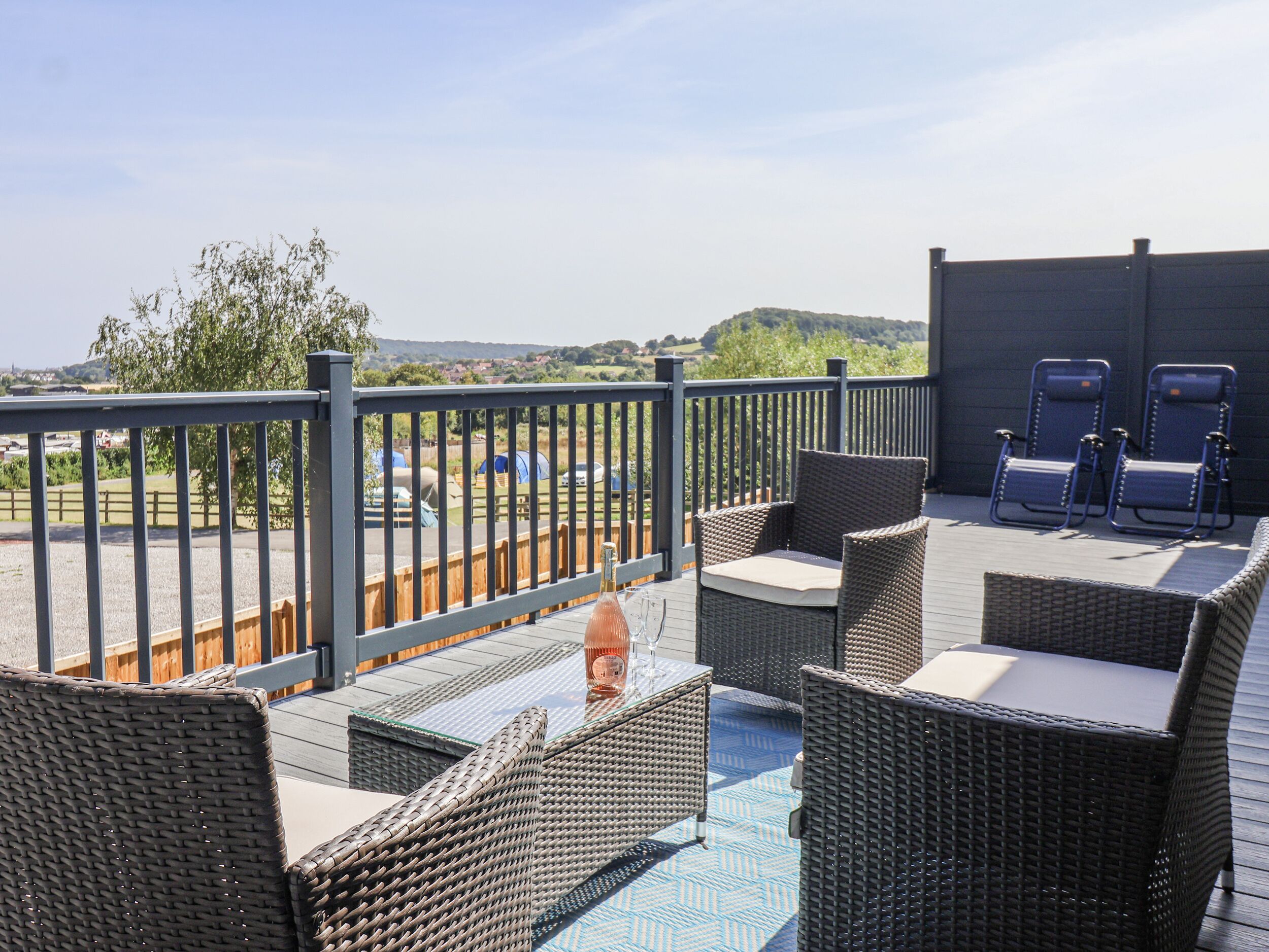 An outdoor deck with wicker furniture, a glass table, and a view of the countryside.
