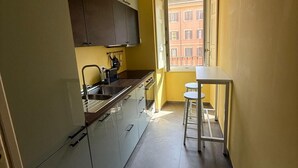 Eine kompakte Küche mit weißen Schränken, ein kleiner Essbereich mit Tisch und zwei Hocker sowie ein Fenster mit Blick auf ein Gebäude.