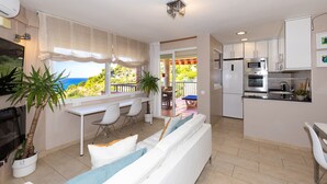 A modern kitchen with white cabinets, a built-in oven, and a microwave. There is a dining area with a white table and chairs, and a large window offering a view of the outdoors.