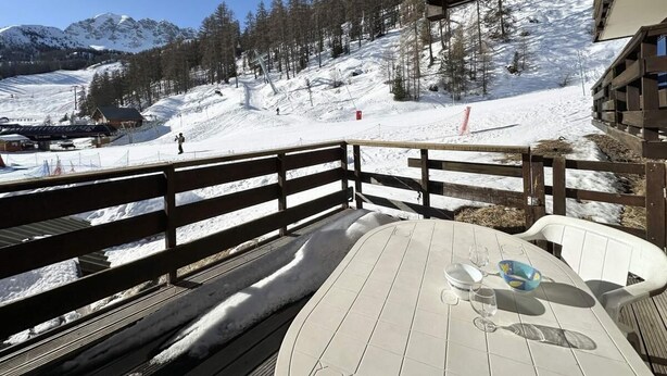A wooden deck with a table and chairs, overlooking a snowy mountain landscape with a ski lift and trees.