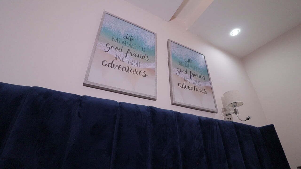 Quarto com cortina azul escura, duas frases inspiradoras emolduradas na parede e uma luminária no teto.