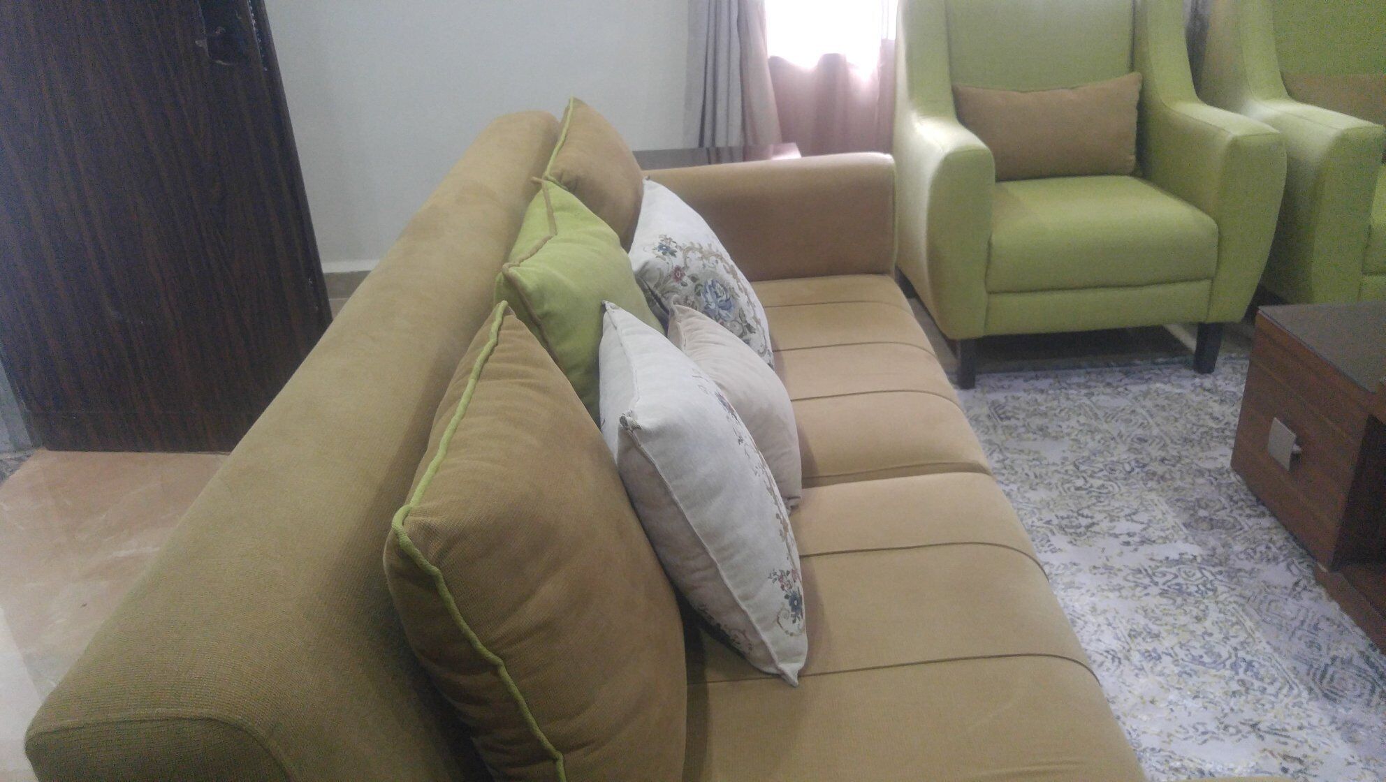 Een woonkamer met een beige bank en groene fauteuils, een houten kast en een kamerbreed tapijt met dessin.