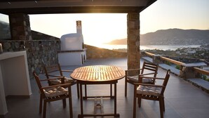 A wooden table with four chairs, a stone wall, and a view of a distant town and water body. - Prime Views A - Peacefull Villa with Private Pool, premium Sea view and Sunsets. (Poseidonia)