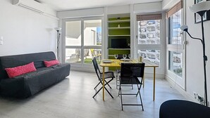Een moderne woonkamer met een bank, eettafel, stoelen en een groot raam met uitzicht op gebouwen.