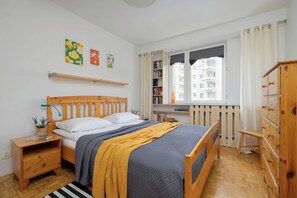 A bedroom with a wooden bed, a bedside table, a nightstand, a window with curtains, a bookshelf, and a dresser. - Spacious Apartment | Two Bedrooms and Balcony (Warszawa)