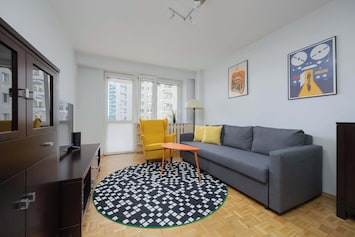 A spacious, bright living room with a grey sofa, yellow armchair, round rug, and TV cabinet. Large windows provide natural light.