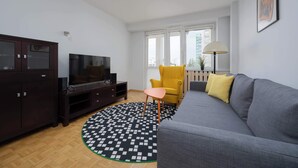 Ein Wohnzimmer mit grauem Sofa, einem gelben Sessel, einem Fernseher auf einem dunklen Holzschrank und einem gemusterten Teppich.