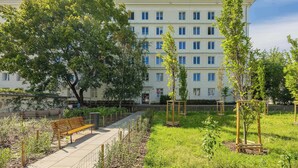 A multi-story building with a garden area featuring a wooden bench, young trees, and a pathway.