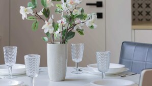 A round dining table with white plates, cutlery, and textured glassware. A white vase with white flowers is placed in the center. The room has a modern design with black leather chairs and a light-colored wall.
