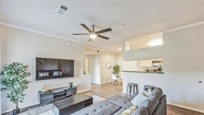A modern living room with a large flat-screen TV, a grey sectional sofa, a ceiling fan, and a kitchen area in the background. - Rocky Mountain Haven w/ king bed, pool & hot tub (Castle Rock)