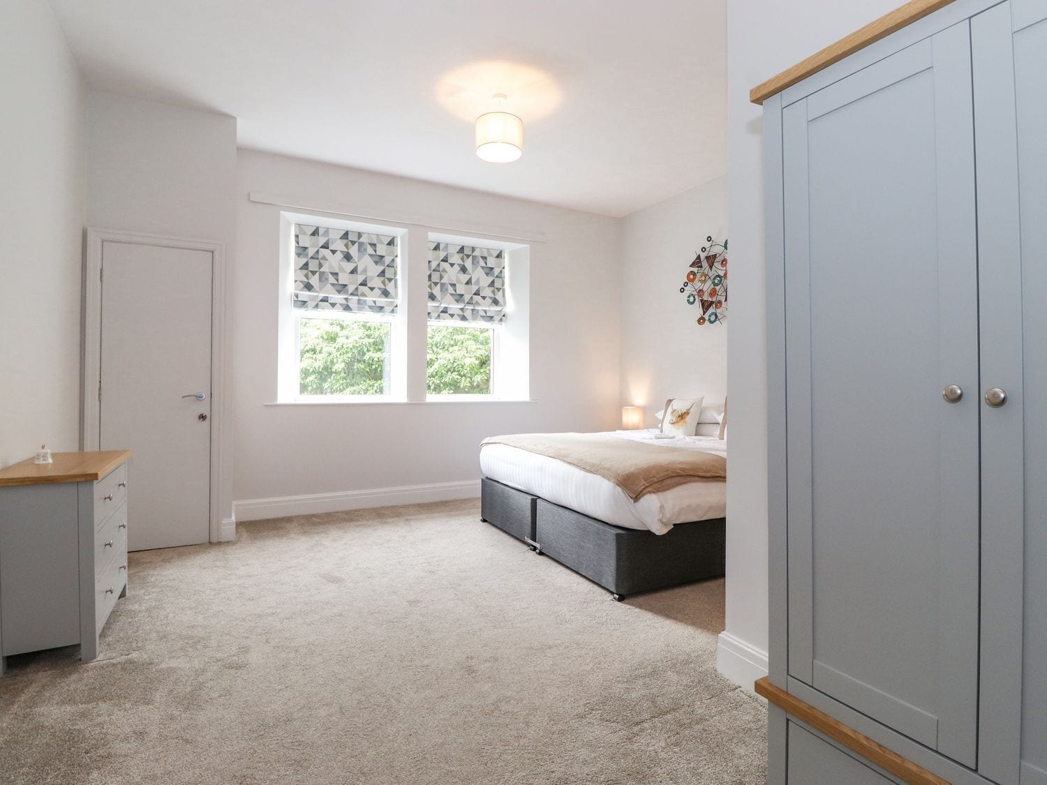 Quarto com cama, guarda-roupa, escrivaninha e duas janelas com cortinas estampadas.