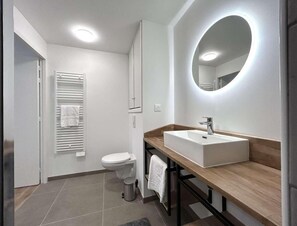 A modern bathroom with a white toilet, a wooden vanity with a sink, and a large mirror with a circular light.