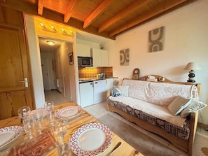 Uma sala de estar aconchegante com sofá, mesa de jantar posta para quatro pessoas e cozinha ao fundo.