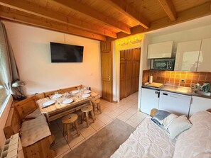 A compact room with a dining table, a bed, a kitchenette, and a television mounted on the wall.