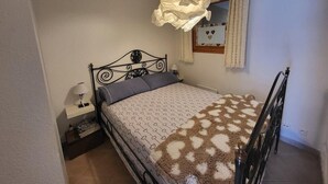 Un dormitorio con cama, mesita de noche, lámpara, ventana con cortinas y aplique de pared.