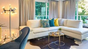 A modern living room with a white sofa, a glass coffee table, and a blue chair. There are curtains on the windows, and a cowhide rug on the floor.