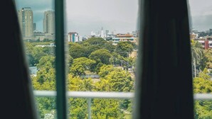 A view through window blinds of a cityscape with dense greenery and high-rise buildings. - Hermoso apartamento con vista a la ciudad (Santo Domingo)