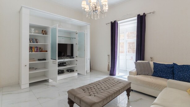 Ein Wohnzimmer mit einem weißen Sofa, einem beigefarbenen Hocker, einem Fernseher auf einem Standfuß und einer weißen Unterhaltungsanlage mit Regalen.