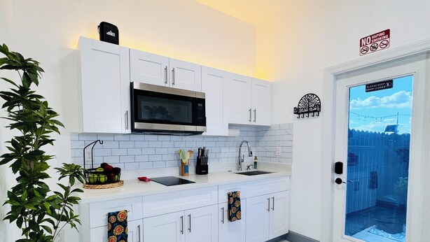 Una cucina moderna con mobili bianchi, un forno a microonde e un lavandino. Sono presenti piante e una porta con il cartello 'No Vaping'.