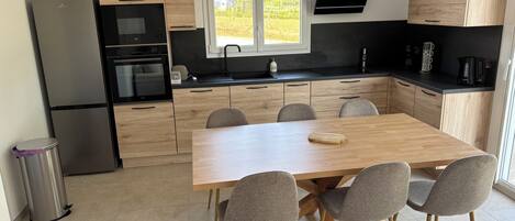 Una cocina moderna con armarios de madera, un refrigerador de acero inoxidable, horno empotrado y comedor con mesa de madera y sillas grises.