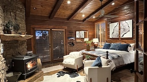 A cozy bedroom with a stone fireplace, a bed with a headboard, and a large window with blinds. - Romantic Winter Escape/Hot tub in Heaven (Pine)